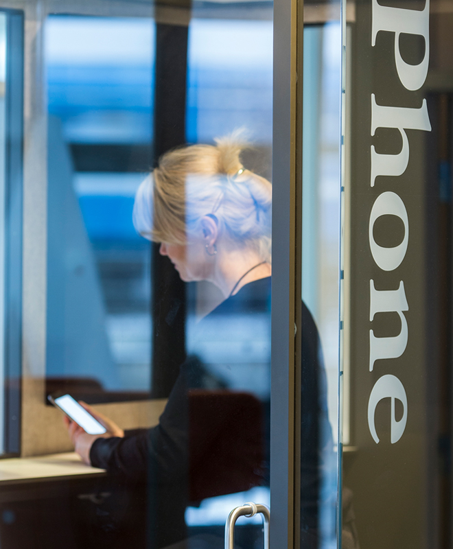 Woman sitting with her phone behind a glass door with a text on it that says " Phone" | Coor 